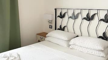 Una cama bien hecha con sábanas y almohadillas blancas, una mesita de noche con lámpara y un aire acondicionado de pared.