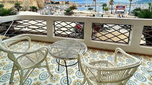 Une terrasse dotée de meubles en osier et d’un sol carrelé aux motifs originaux, offrant une vue sur une marina avec des bateaux et une ville située au loin.