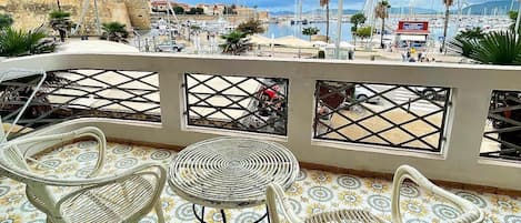 A terrace with wicker furniture and patterned floor tiles, overlooking a marina with boats and a distant town.