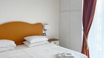 Een net opgemaakt bed met witte lakens, een houten hoofdbord, een nachtkastje met een lamp en een rode kroonluchter.