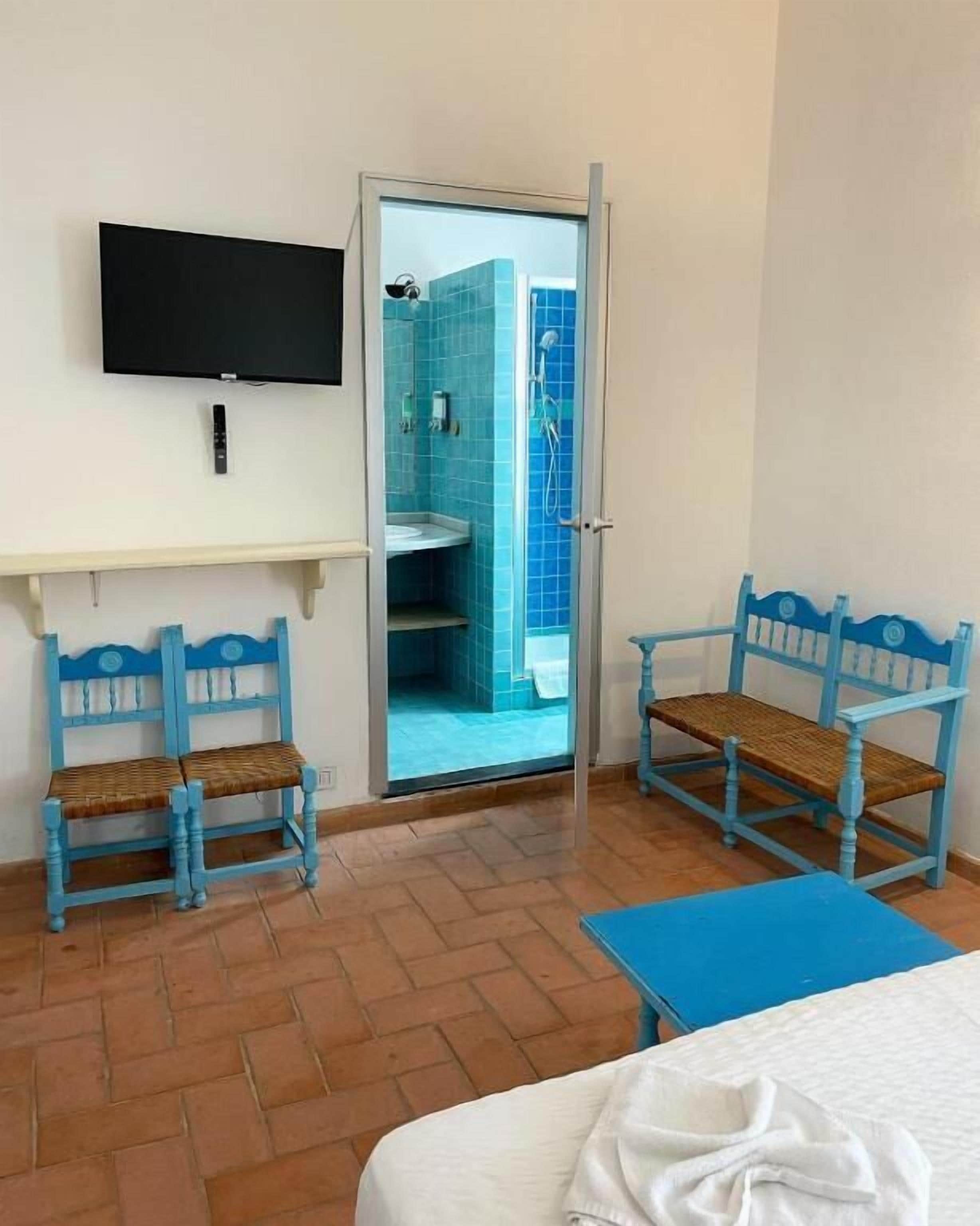 A room with a bed, a TV mounted on the wall, two blue chairs, a small table, and a bathroom visible through an open door.