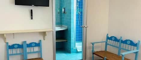A room with a bed, a TV mounted on the wall, two blue chairs, a small table, and a bathroom visible through an open door.