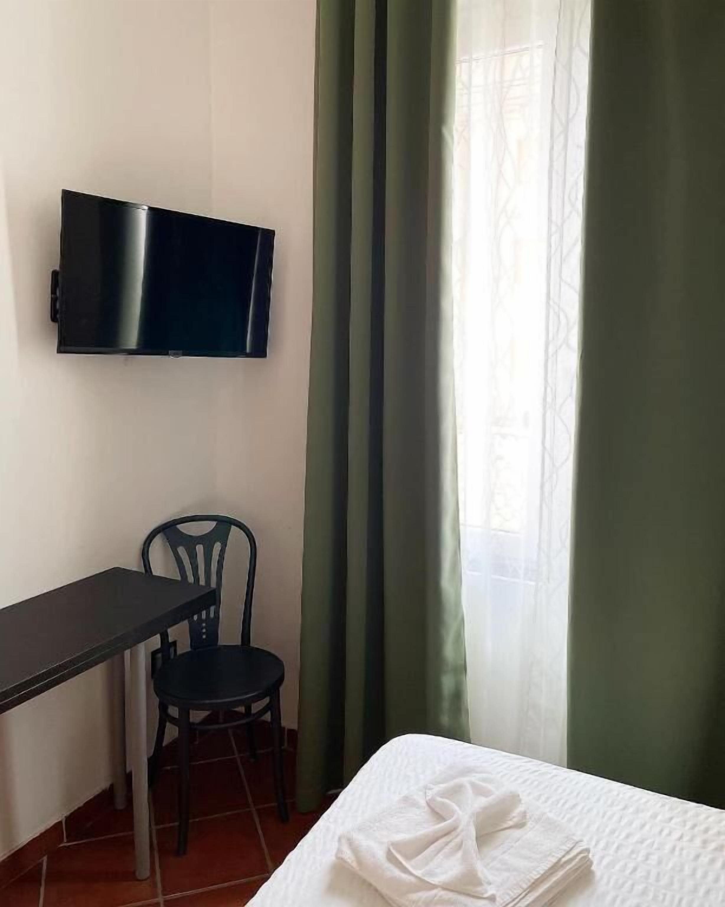 A room with a bed, a desk, a chair, a television mounted on the wall, and a window with curtains.
