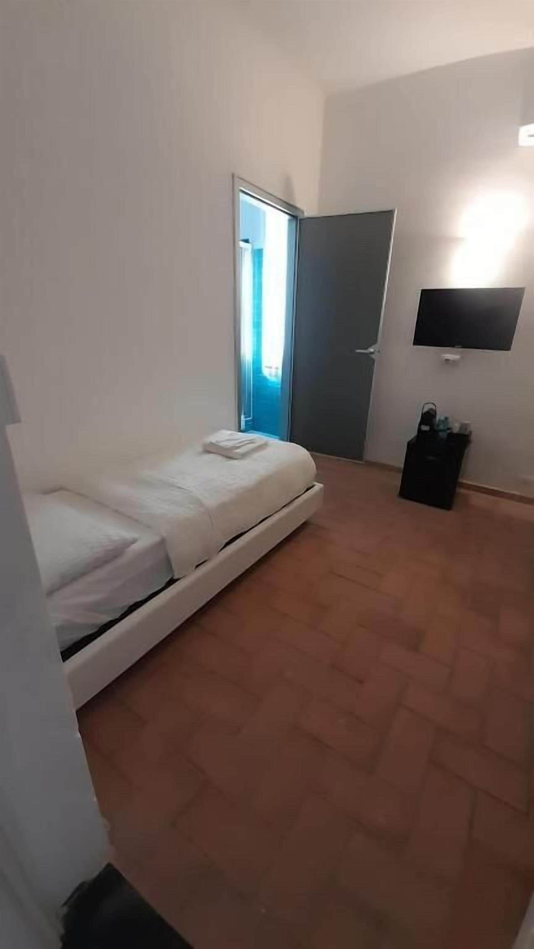 A small, simple room with a bed, a door, a wall-mounted TV, and a small trash bin.