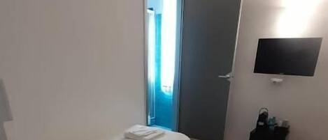 A small, simple room with a bed, a door, a wall-mounted TV, and a small trash bin.