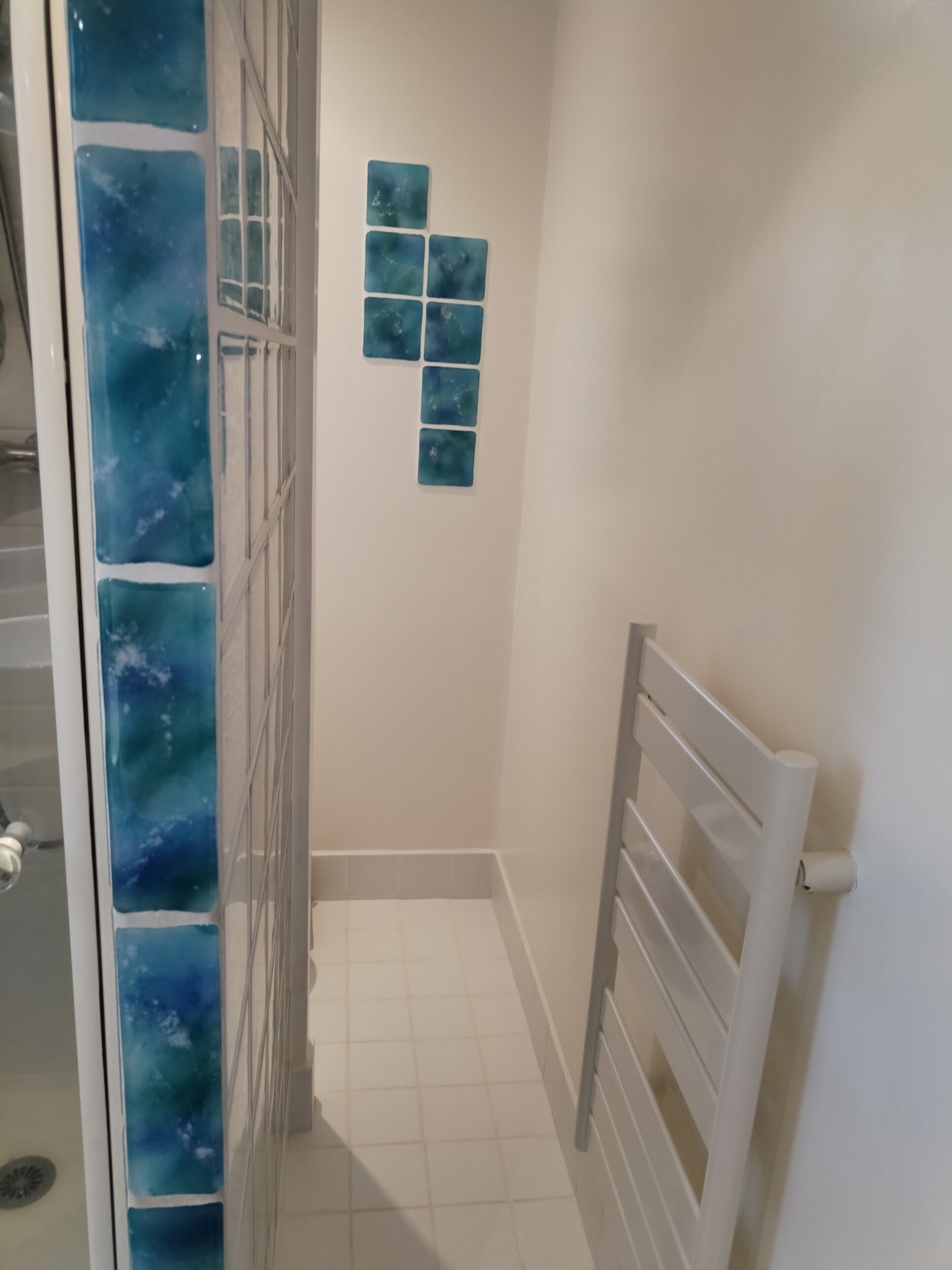 A bathroom with a shower, white tiles, a glass door with a blue pattern, and a white towel rack.