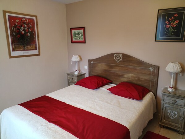 Un lit bien fait, agrémenté d’oreillers rouges, une tête de lit en bois ornée d’un motif en forme de cœur, une table de chevet avec une lampe, et deux tableaux encadrés accrochés au mur.