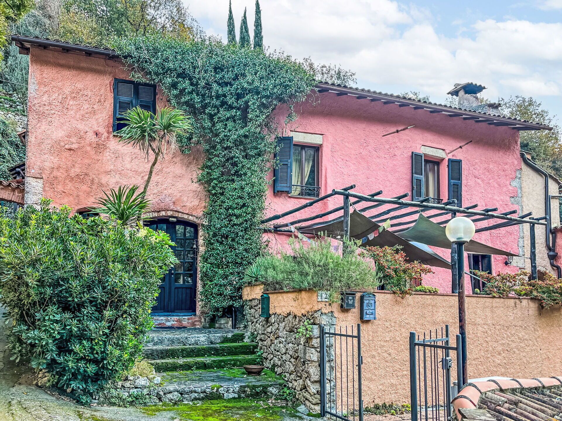 A pink building with a stone staircase leading to a black door, surrounded by greenery and a metal gate.