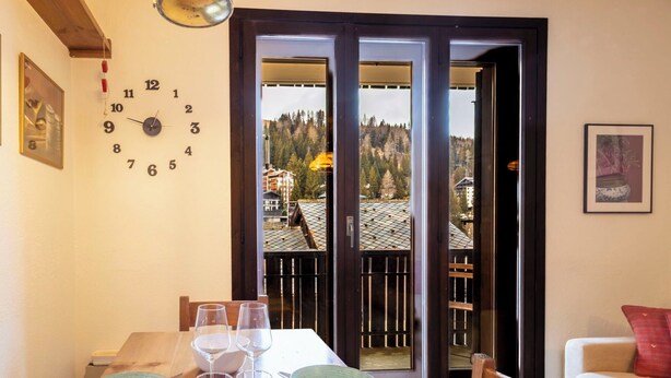 Ein Essbereich mit einem Holztisch, gedeckt für vier Personen, einer Wanduhr und Blick auf eine Berglandschaft durch eine Glastür.