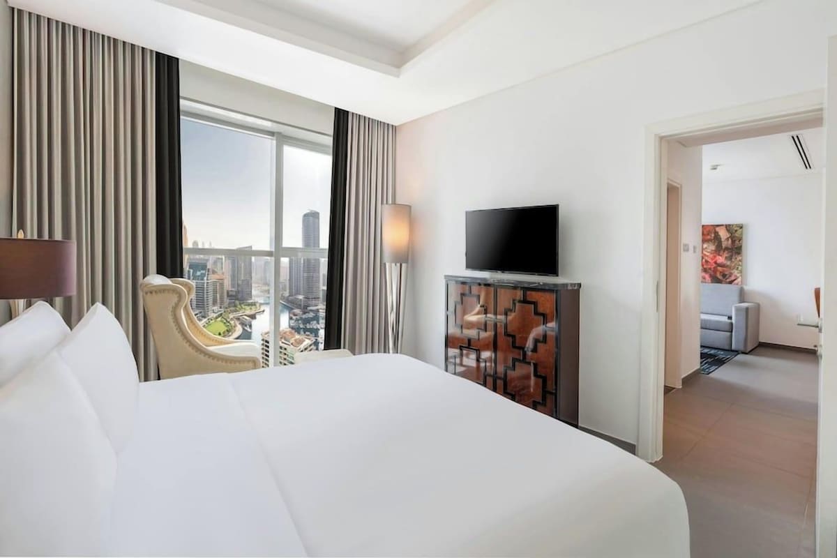 A hotel room with a large bed, a television on a cabinet, a view of the cityscape, and a door leading to another room.