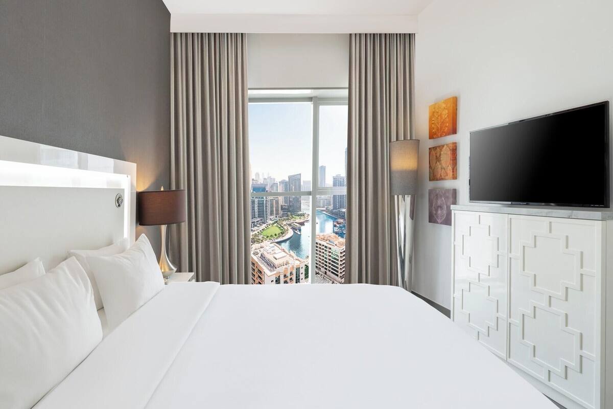 A hotel room with a large bed, a TV, a view of a cityscape, and a window with curtains.