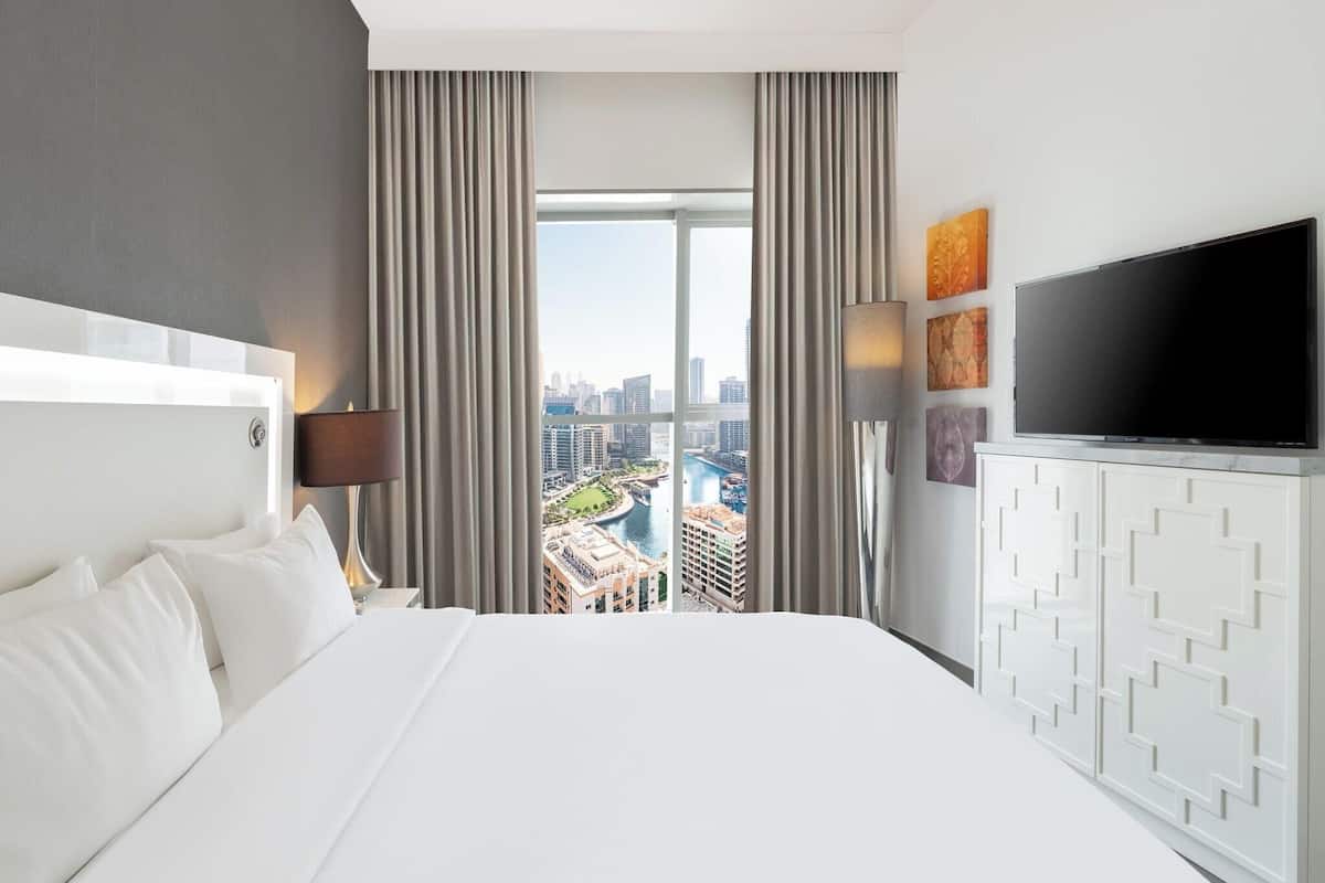 A hotel room with a large bed, a TV, a view of a cityscape, and a window with curtains.