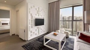 A modern hotel room with a large window, a flat-screen TV mounted on a white geometric wall, a sofa, a coffee table, and a small side table. - The First Collection Marina (Dubai)