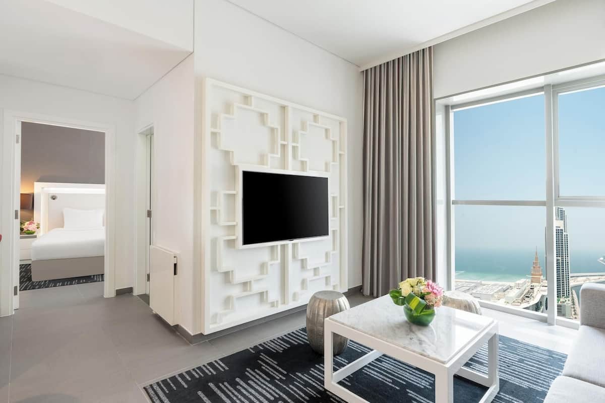 A modern hotel room with a large TV, a bed, a sofa, a view of the city, and a decorative wall panel.