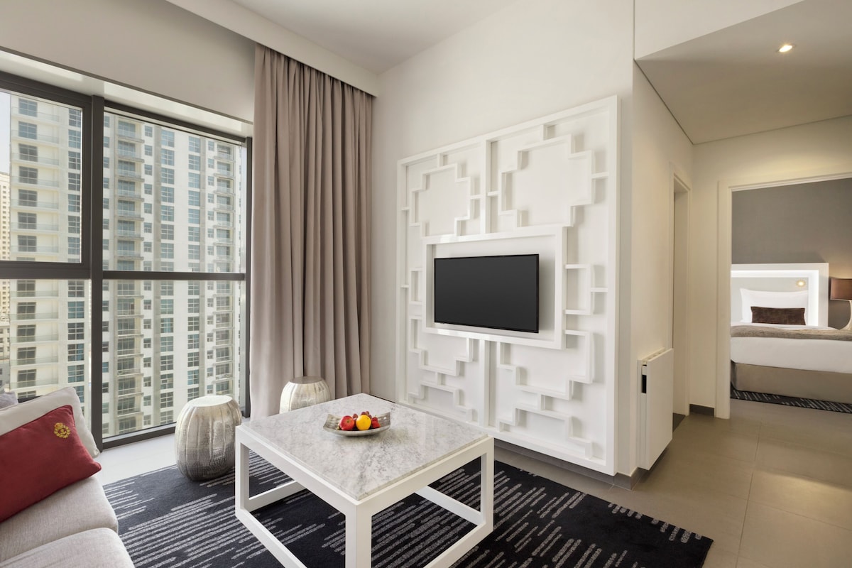 Habitació d'hotel moderna amb un finestral gran, un televisor de pantalla plana al mur blanc, una taula de café de marbre i un llit amb capçalera.