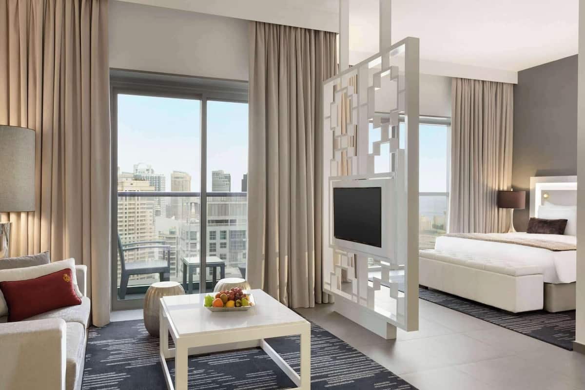 A modern hotel room with a large window, a flat-screen TV, a bed, a sofa, and a coffee table with fruit.