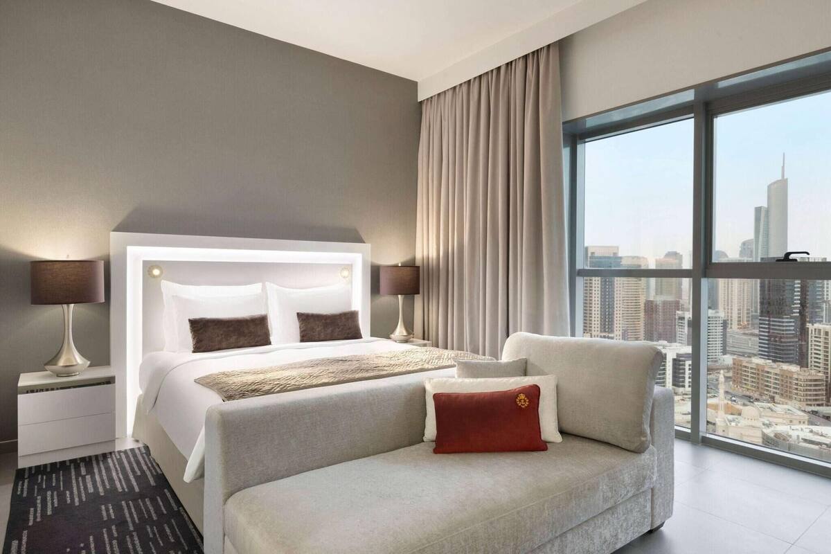 A modern hotel room with a large bed, a sofa, a nightstand, and a view of a cityscape through large windows.