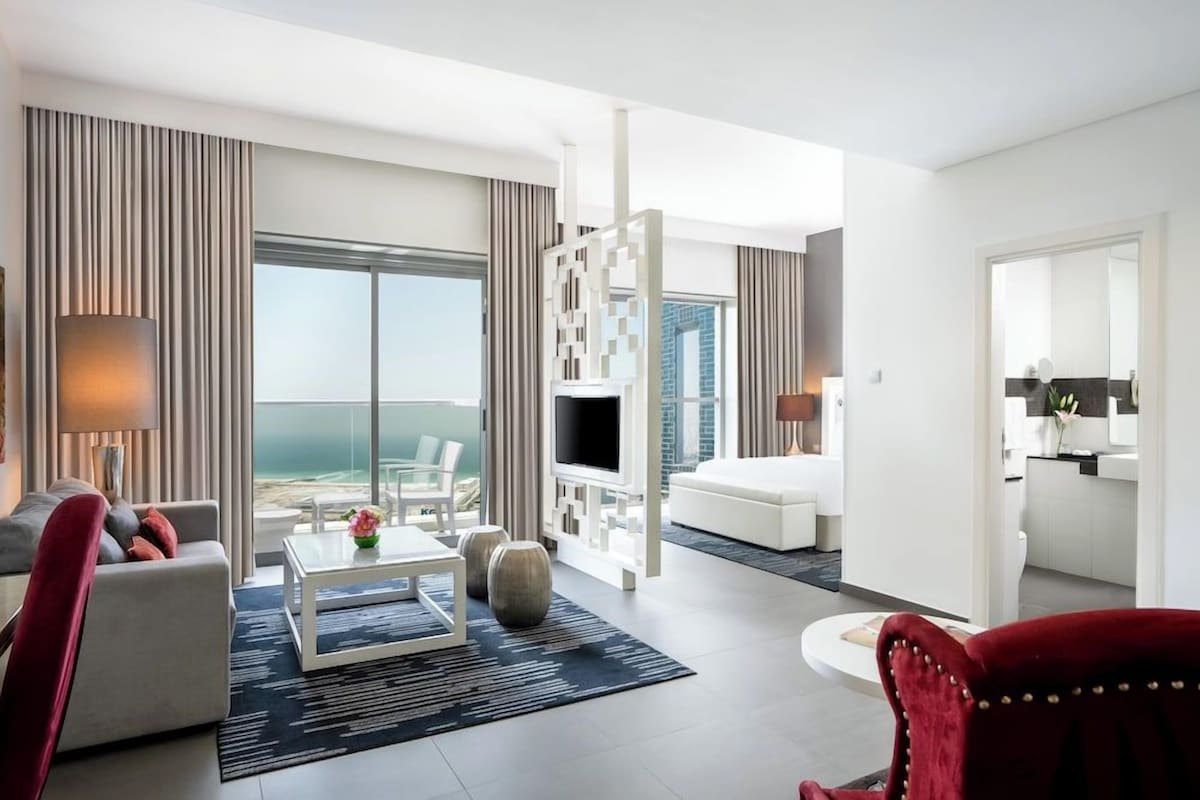 A modern hotel room with a balcony, a flat-screen TV, a sofa, a coffee table, and a red armchair.