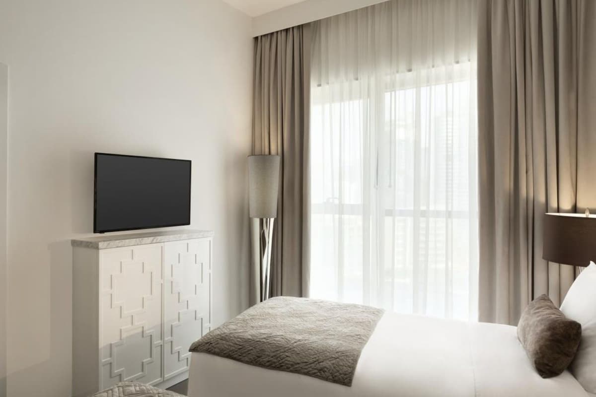 A hotel room with a bed, a TV on a white cabinet, a window with curtains, and a lamp.