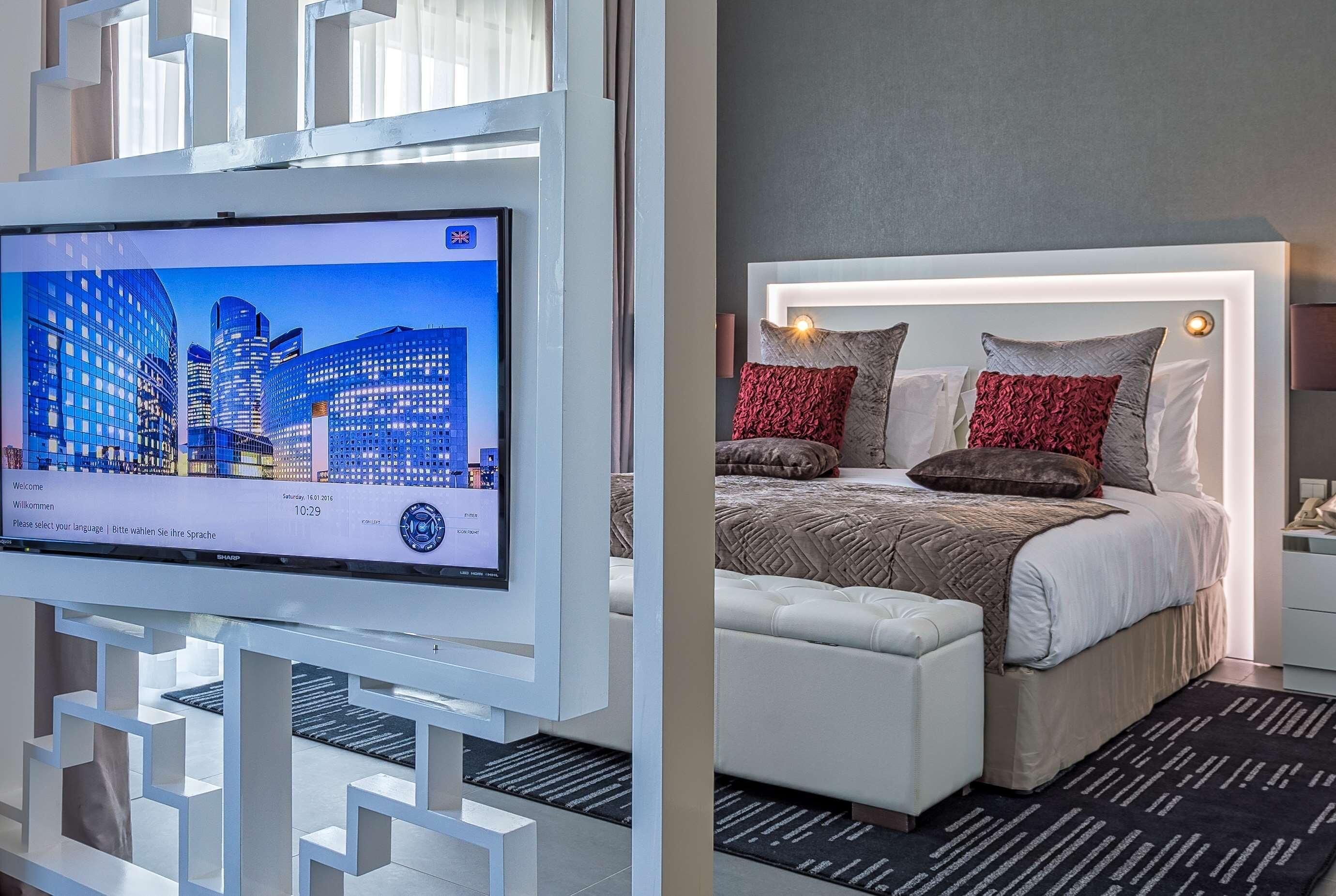 A modern hotel room with a large flat-screen TV, a bed with a white headboard, and a patterned rug.