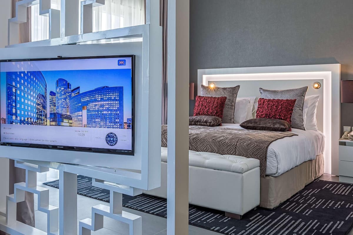 A modern hotel room with a large flat-screen TV, a bed with a white headboard, and a patterned rug.