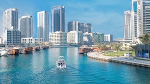 A waterfront cityscape with high-rise buildings, a marina with boats, and a promenade with palm trees. - The First Collection Marina (Dubai)