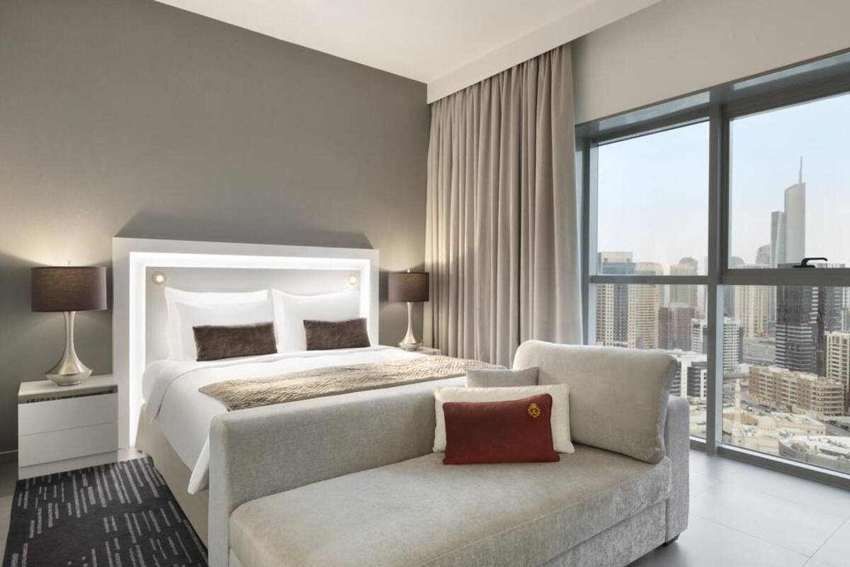 A modern hotel room with a large bed, a sofa, a nightstand, a lamp, and a view of the city.