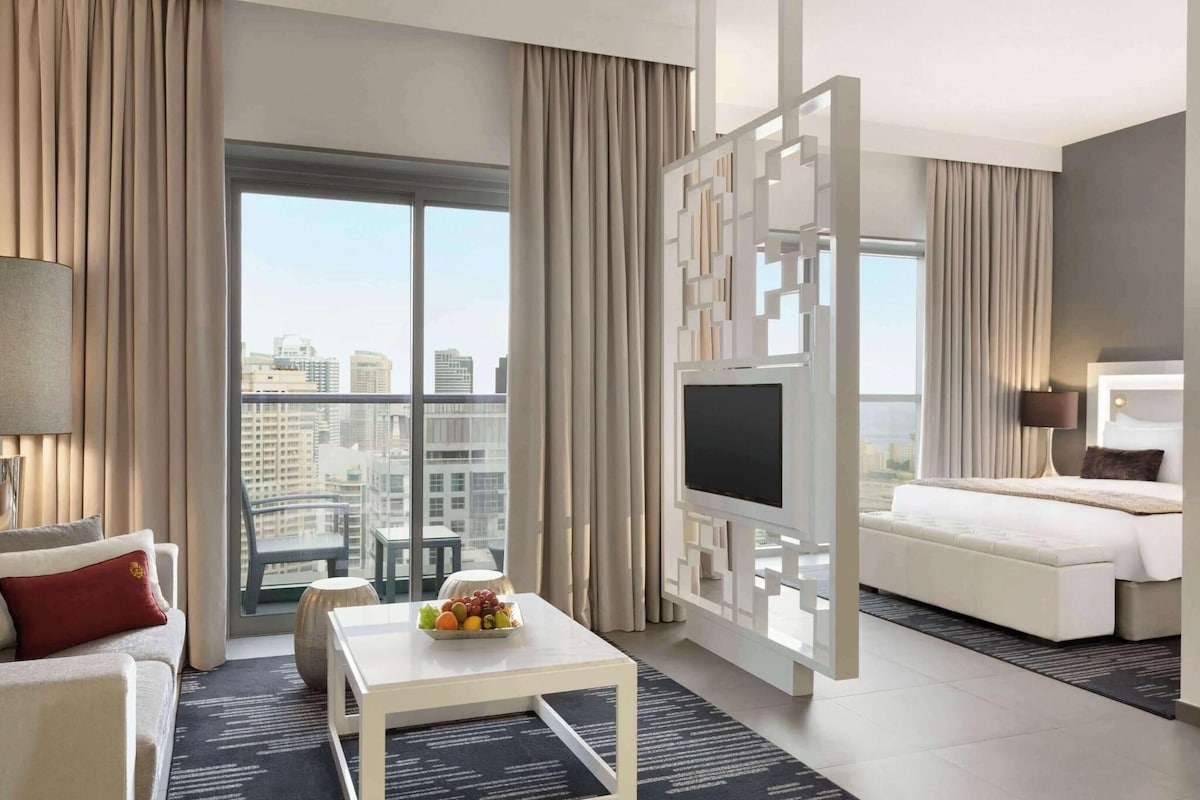 A modern hotel room with a large window, a flat-screen TV, a bed, a sofa, and a coffee table with fruit.
