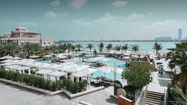 A resort pool area with multiple lounge chairs and umbrellas, a sign for 'Soluna Beach Club,' and a view of the sea and city skyline. - The First Collection Marina (Dubai)