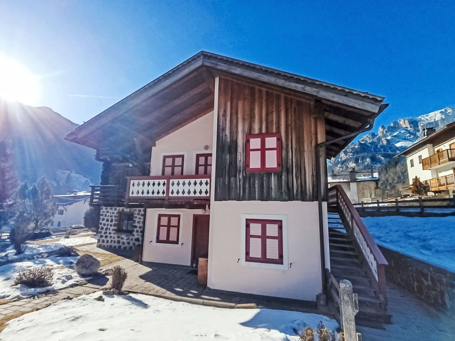 A two-story wooden chalet with a balcony, set against a snowy mountain backdrop.