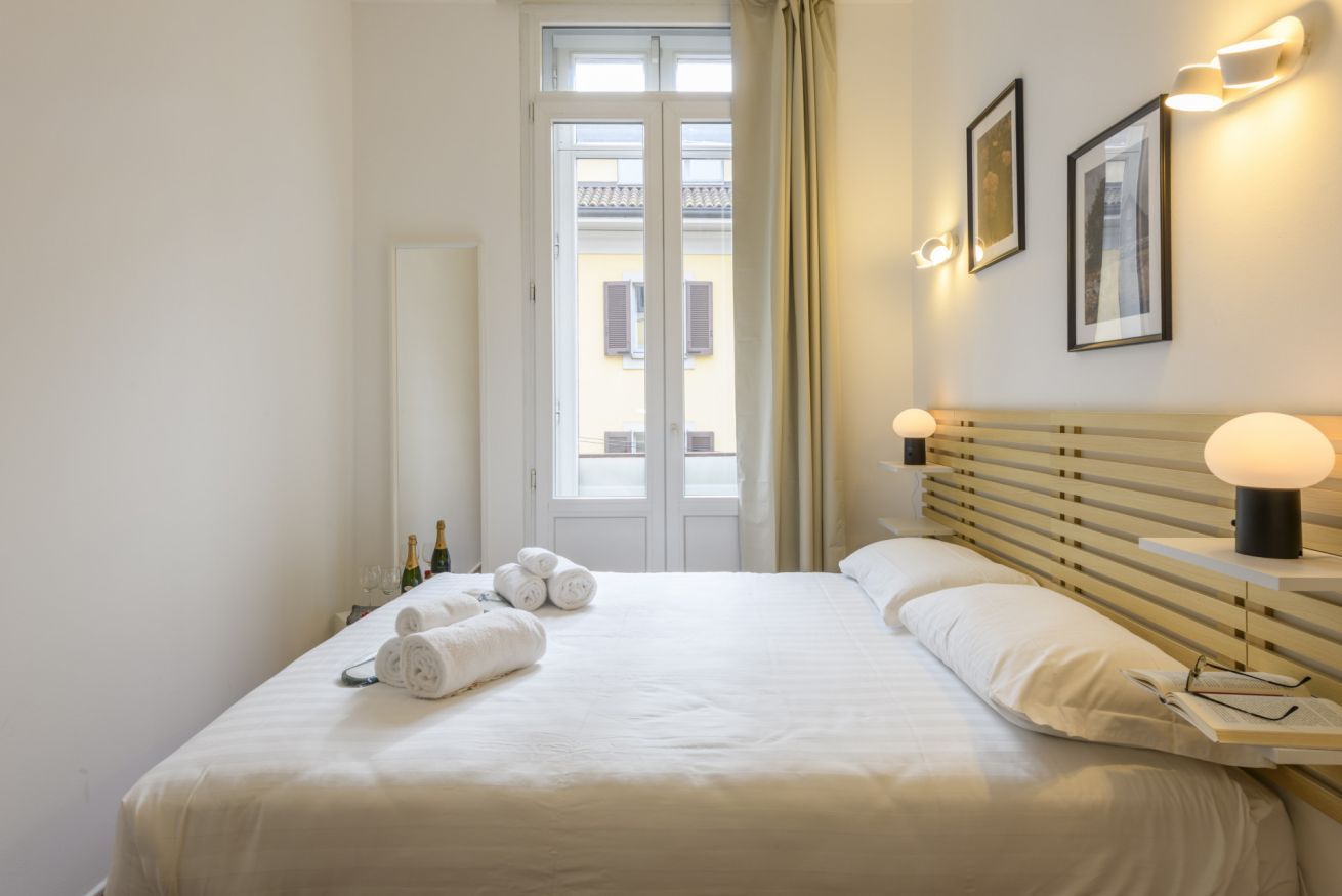A hotel room with a bed, two bedside lamps, a window with curtains, and a door.