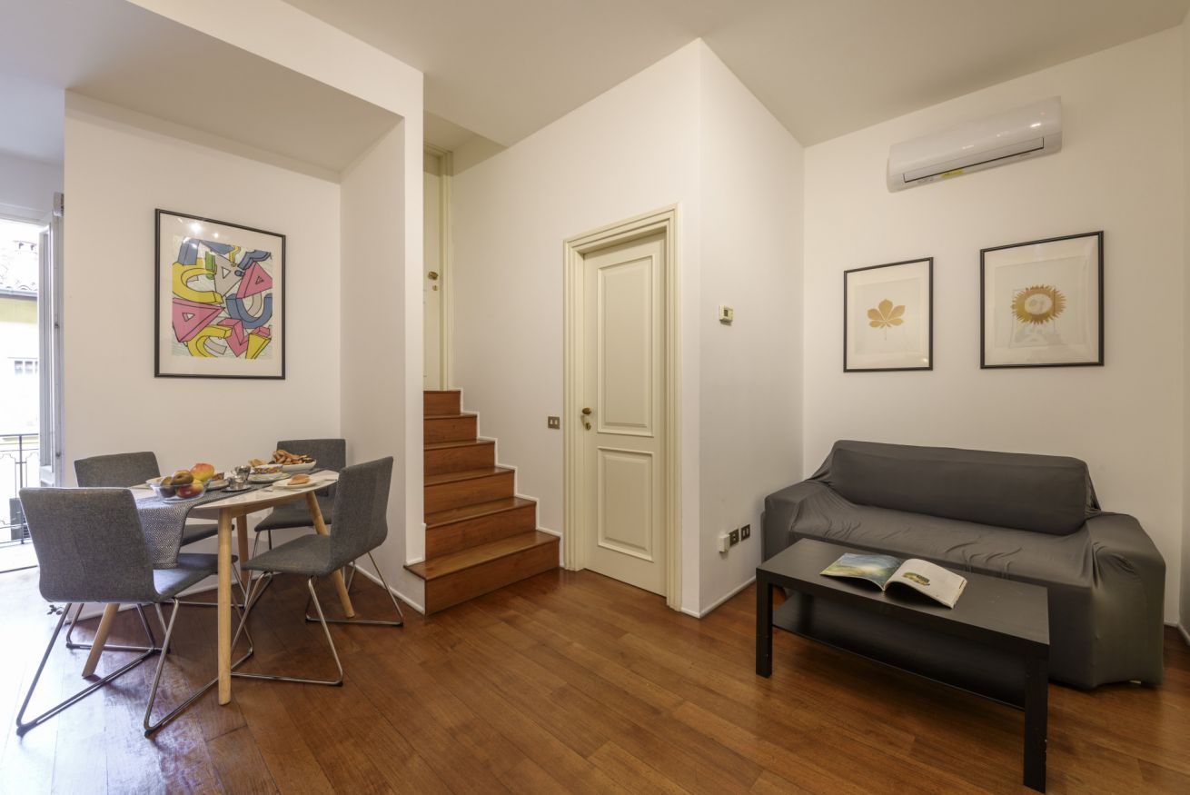A modern living room with a gray sofa, a dining table with chairs, a staircase, and a door.