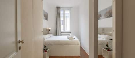 Un dormitorio con cama, mesitas de noche, una ventana y una puerta.