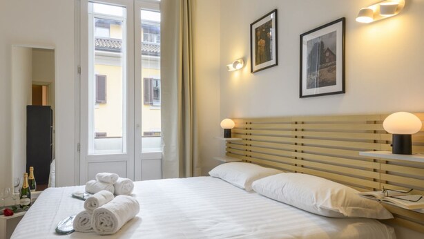 A hotel room with a bed, two bedside lamps, a nightstand with a bottle and glasses, and a window with a view of buildings. - PONTACCIO, 4 - LIVING IN BRERA, IN THE HEART OF MILAN (Milan)