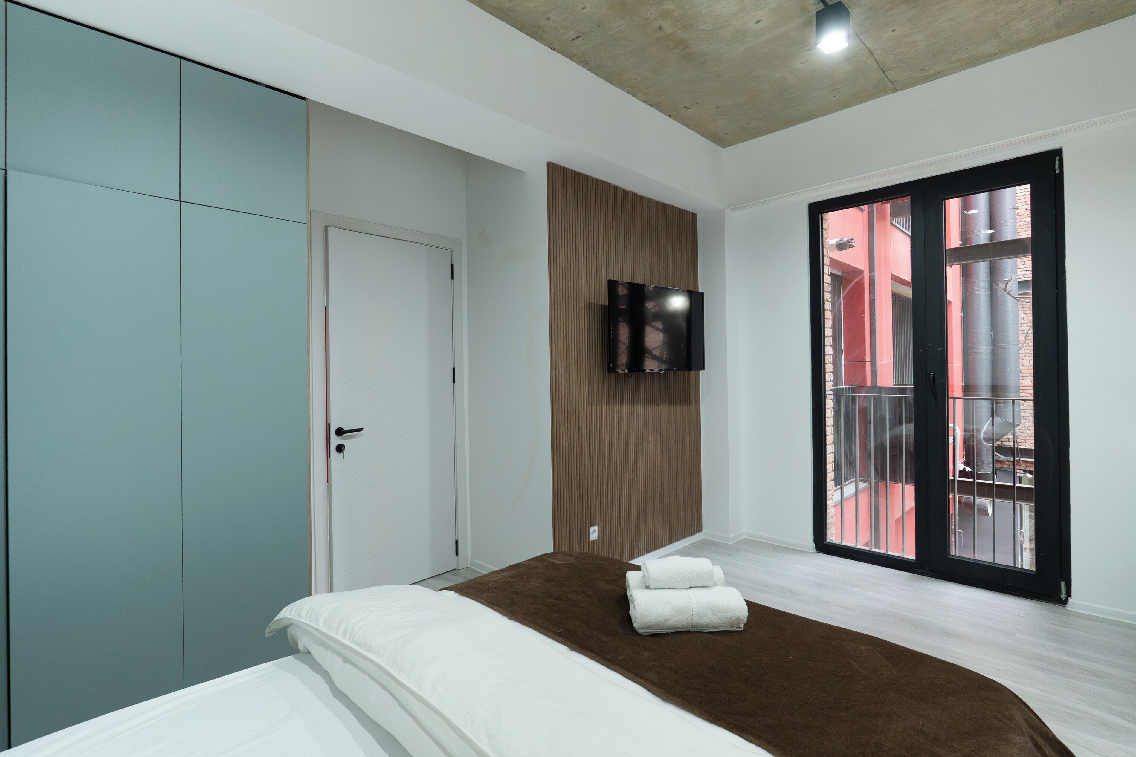 Un dormitorio moderno con cama, armario, televisión y balcón con barandillas rojas.