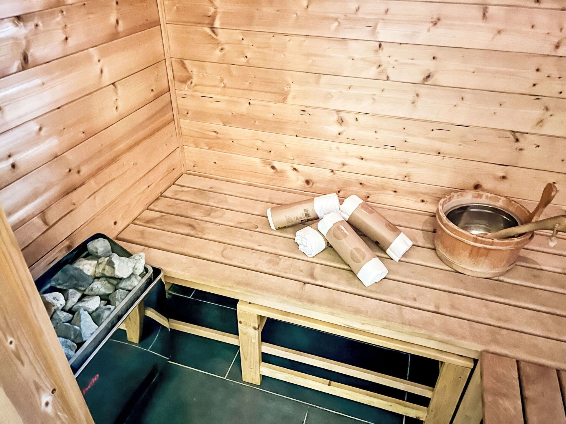 A sauna room with wooden walls, a bench, a bucket, and a container of stones.