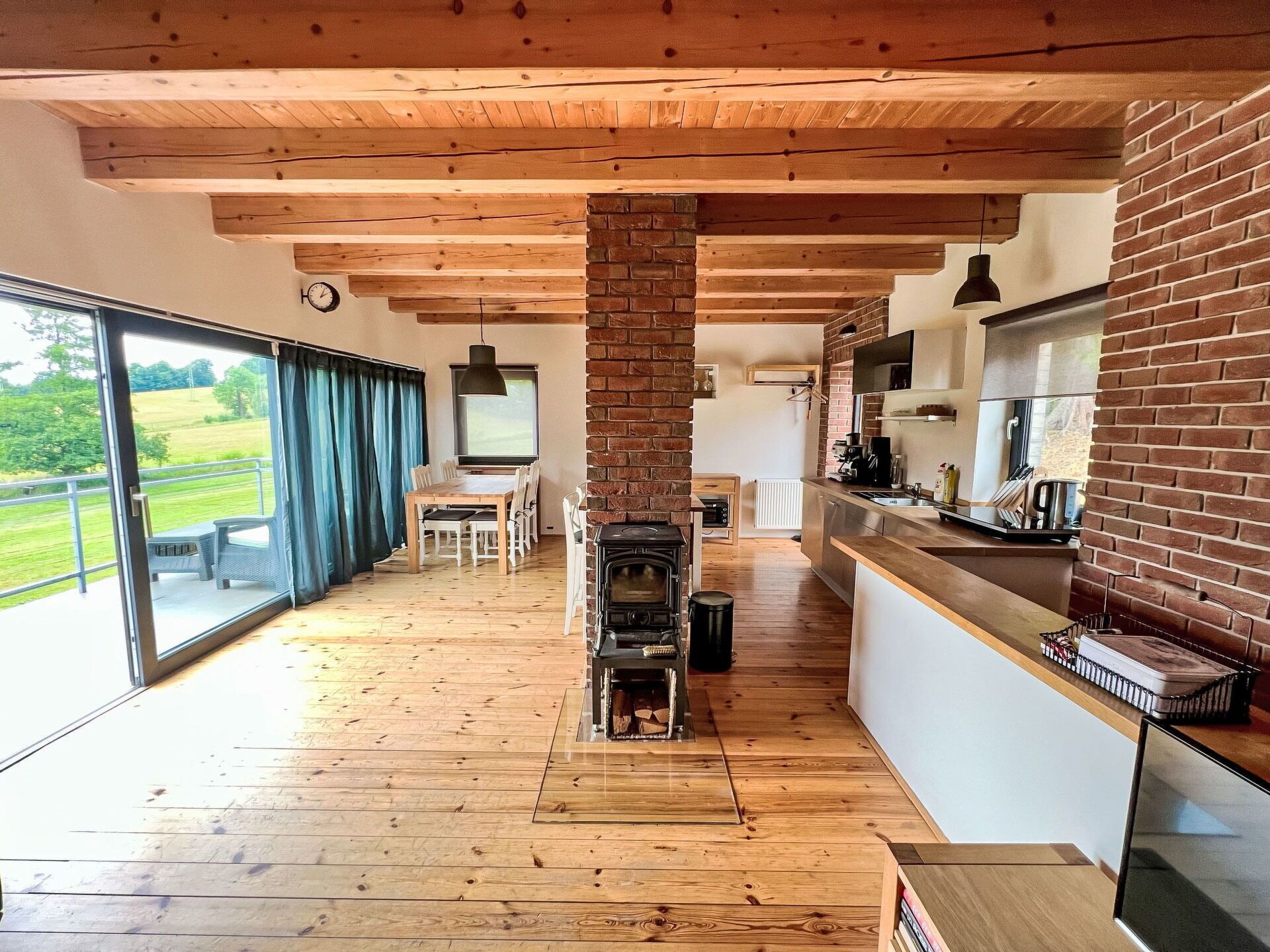 Une cuisine moderne avec une colonne centrale en briques, des poutres en bois, un coin repas avec une table et des chaises, et une grande porte-fenêtre coulissante donnant sur un balcon.