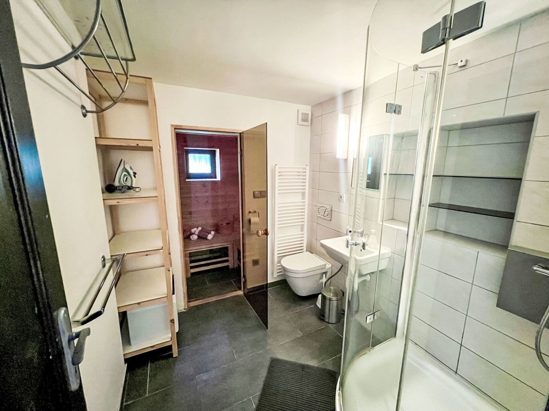 A modern bathroom with a glass shower enclosure, a toilet, a sink, and a small shelf with toiletries.