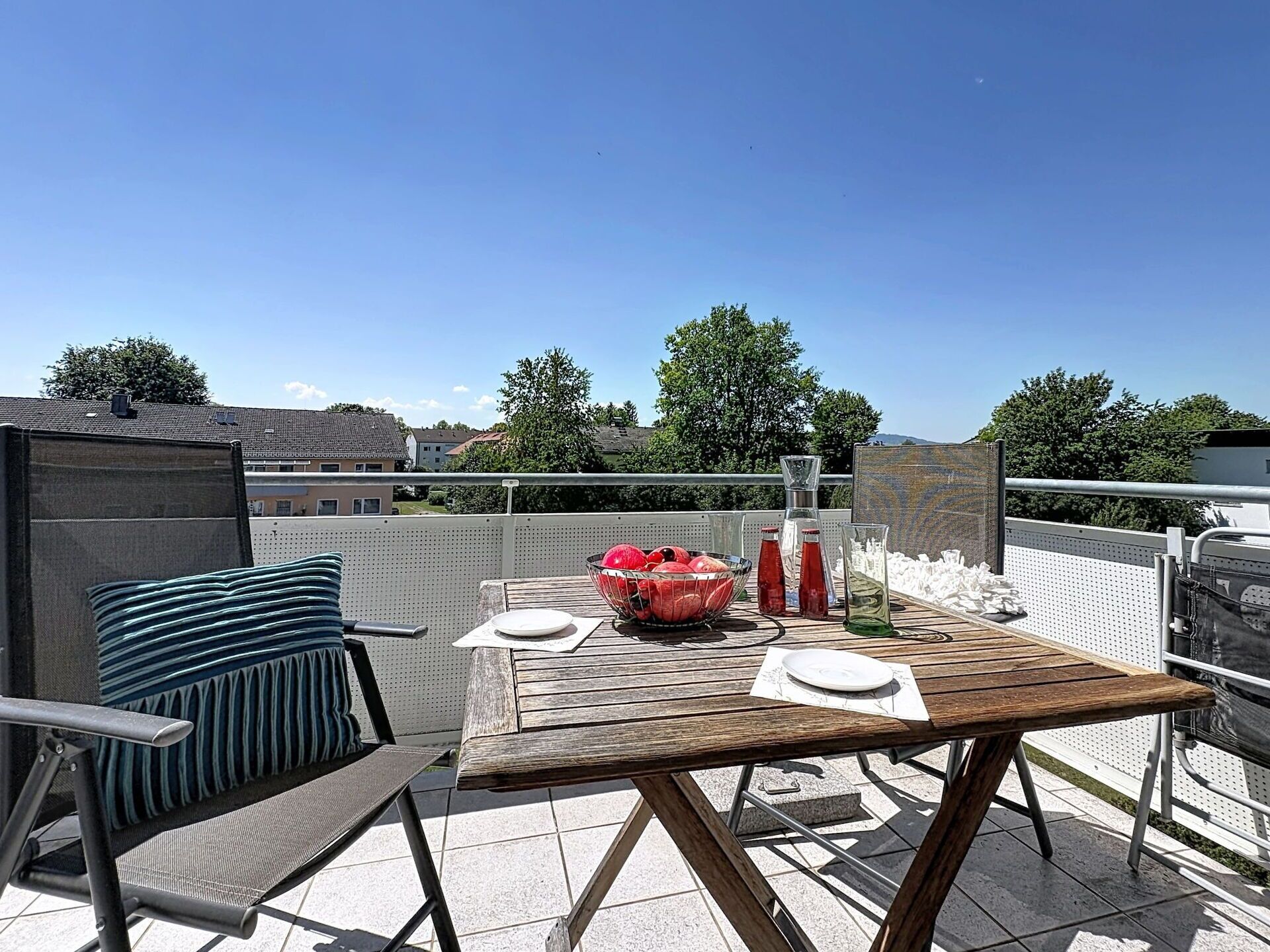 Een houten tafel met een witte tafelkleed, een schaal met fruit en twee flessen op een balkon met uitzicht op huizen en bomen.
