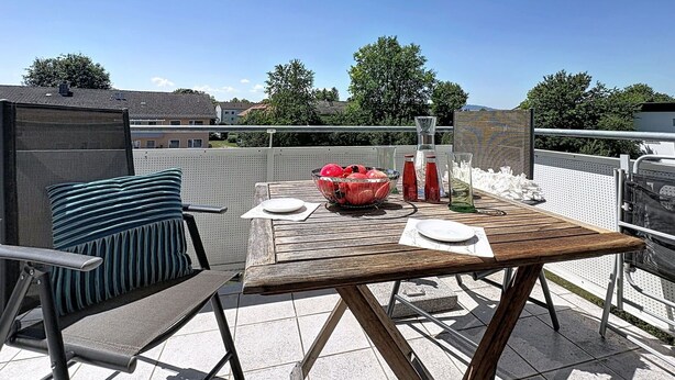 Ein Holztisch mit weißem Tischtuch, eine Obstschale und zwei Flaschen auf einem Balkon mit Blick auf Häuser und Bäume.