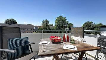 Ein Holztisch mit weißem Tischtuch, eine Obstschale und zwei Flaschen auf einem Balkon mit Blick auf Häuser und Bäume.