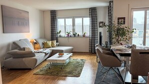 A modern living room with a sofa, coffee table, and dining area. There are windows with curtains, a plant, and a painting on the wall.