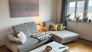 A modern living room with a grey sectional sofa, a glass coffee table with fruits, a remote control, and a lamp.