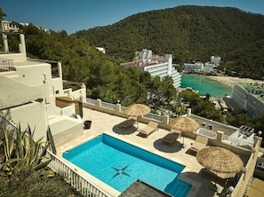 A rooftop pool with lounge chairs and umbrellas, surrounded by a mountainous landscape and a beach.