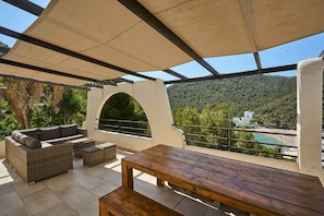 Eine Terrass mit Holztable und Rattan-Sofa mit Blick auf einen Strand und bewaldete Hügel.