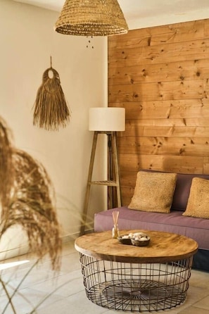 Ein Zimmer mit einer Holzwand, einem violetten Sofa, einem runden Couchtisch mit Webdecke, einer Stehlampe und einer hängenden Makramee-Leuchte.