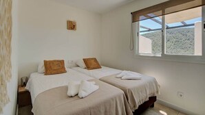 Quarto com duas camas, cada uma com colcha branca e travesseiro marrom. Uma pequena estante de madeira com uma luminária está do lado esquerdo. Uma janela com vista para uma área arborizada fica do lado direito.