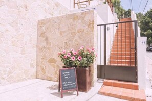 A stone wall, a flower planter with pink flowers, a signboard with text, and a staircase with a metal railing. - Apartment nº7, 1 bedroom, 1 bathroom (Cala Llonga)