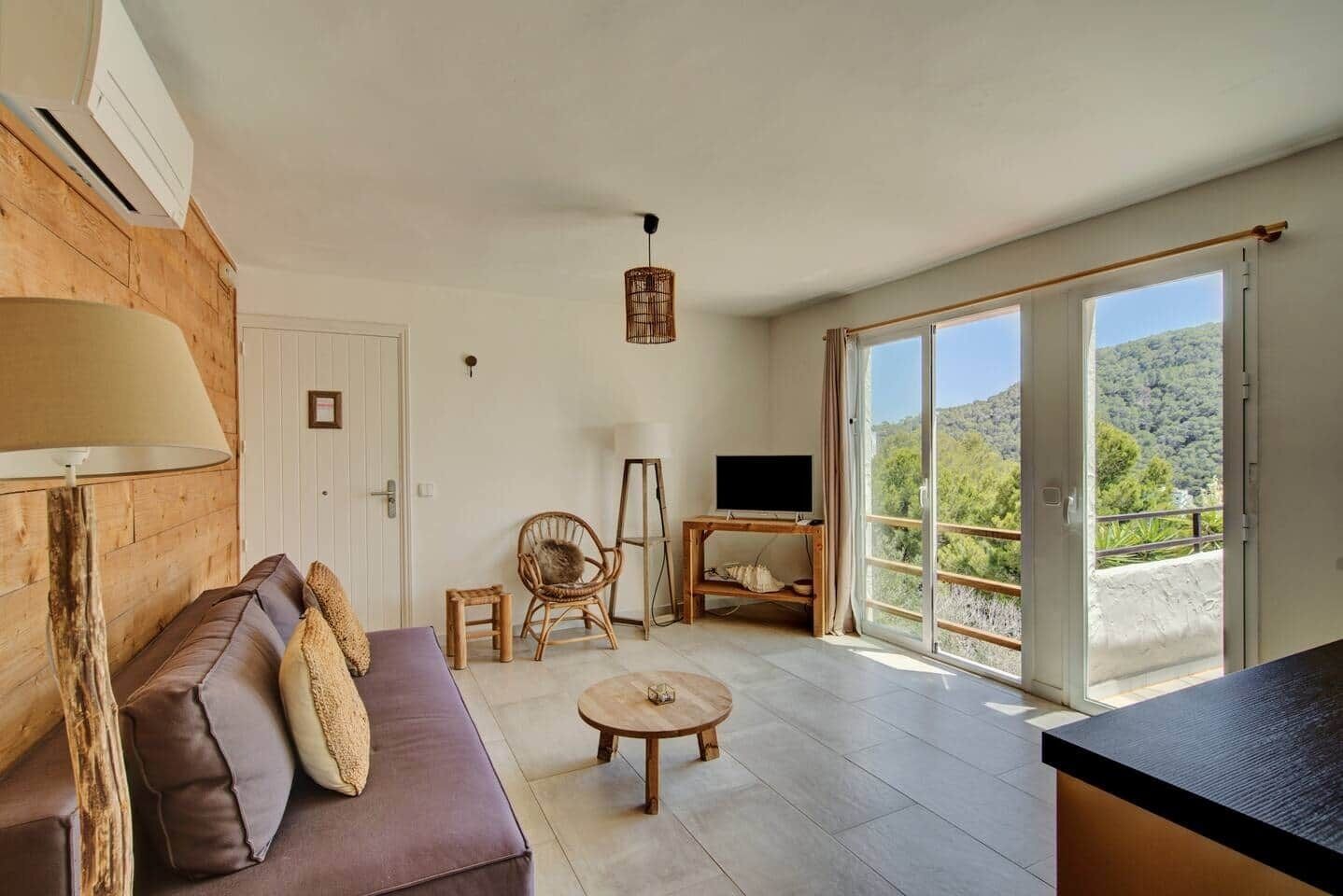 Een moderne woonkamer met een bank, een lamp, een televisie op een standaard, en een grote schuifglazen deur die leidt naar een balkon met een prachtig uitzicht.