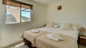 A bedroom with two beds, a window with a view of a hill, a small wall-mounted light, and a wooden nightstand. - Apartment nº7, 1 bedroom, 1 bathroom (Cala Llonga)